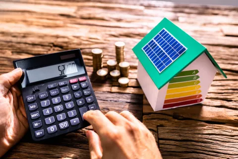 Zwei Hände tippen auf einem Taschenrechner, während daneben ein kleines Modell eines Solarhauses steht und ein Stapel Geldmünzen.